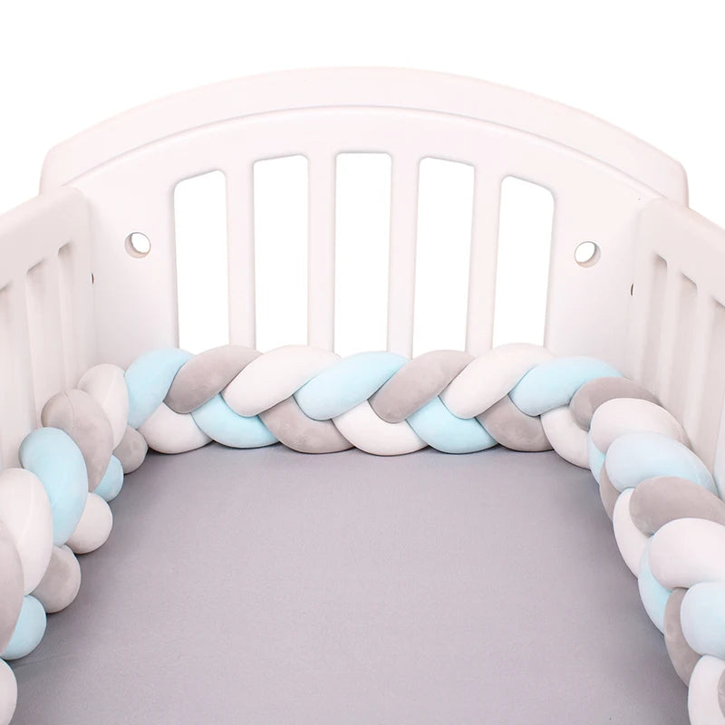 White crib featuring a braided bumper in pastel blue, gray, and white colors, surrounding the edges of a gray mattress, offering softness and protection for a baby.