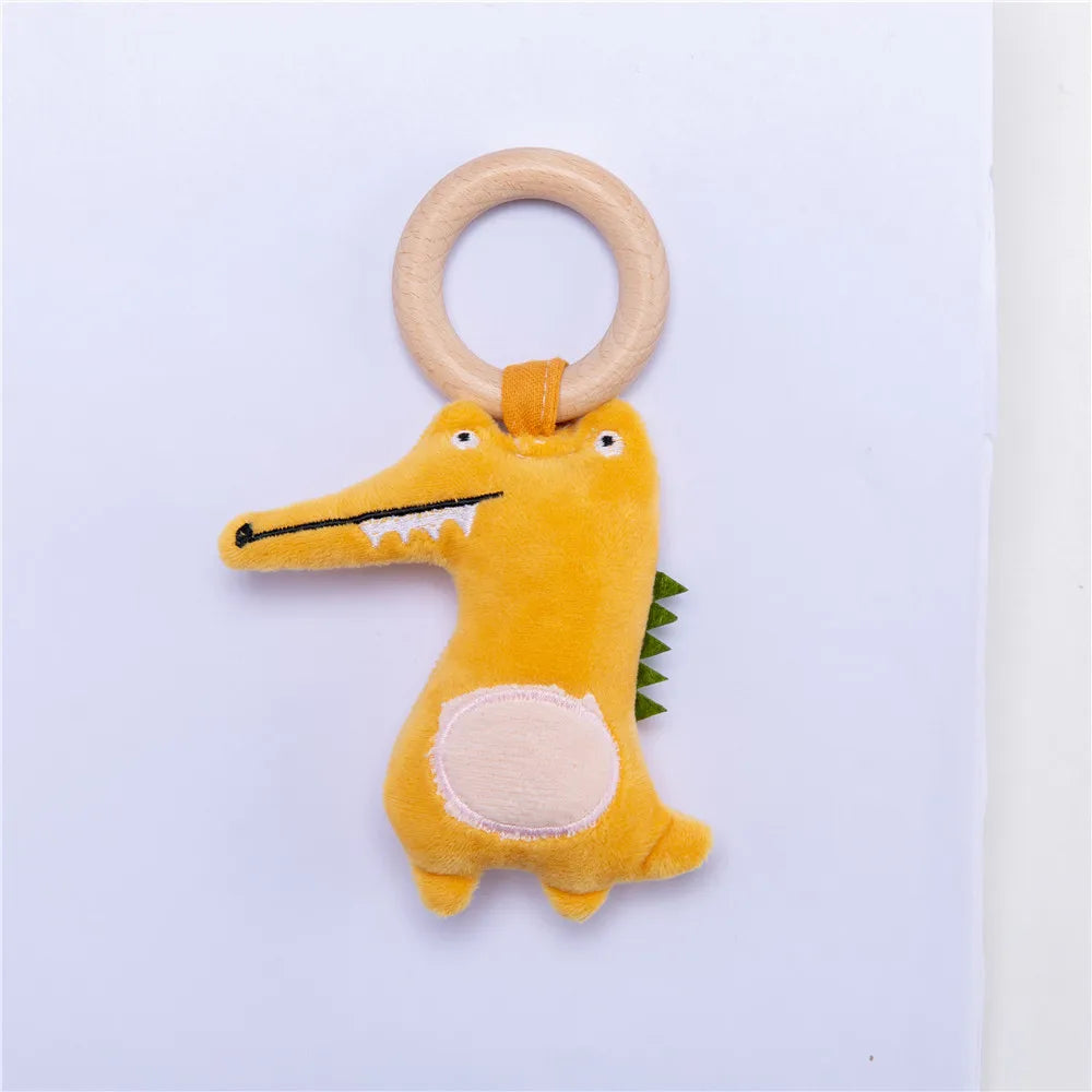 A plush yellow crocodile-shaped toy with a wooden ring attached. The toy has white eyes and a stitched mouth with teeth, along with green felt spikes on its back.