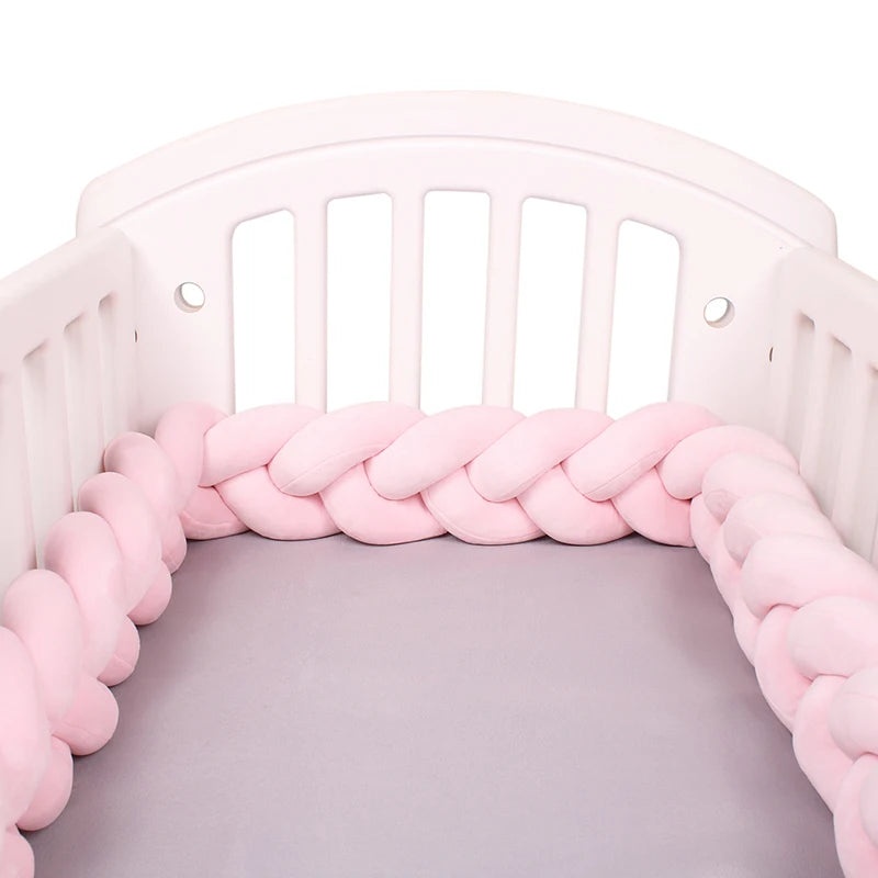 A white baby's crib with a padded pink braided bumper lining the interior, sitting atop a soft gray mattress.