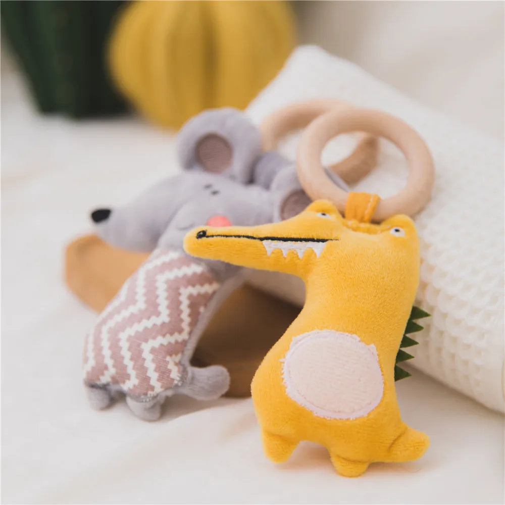 Soft plush toys on a white surface, featuring a yellow crocodile with a wooden ring and a gray mouse with a zigzag pattern. A blurred yellow object is in the background.