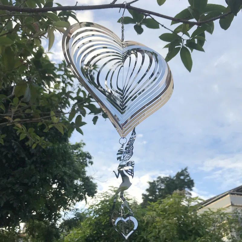 A metallic heart-shaped wind spinner hangs from a tree branch against a blue sky. Its layered design creates a mesmerizing effect, with dangling elements adding to its elegance.