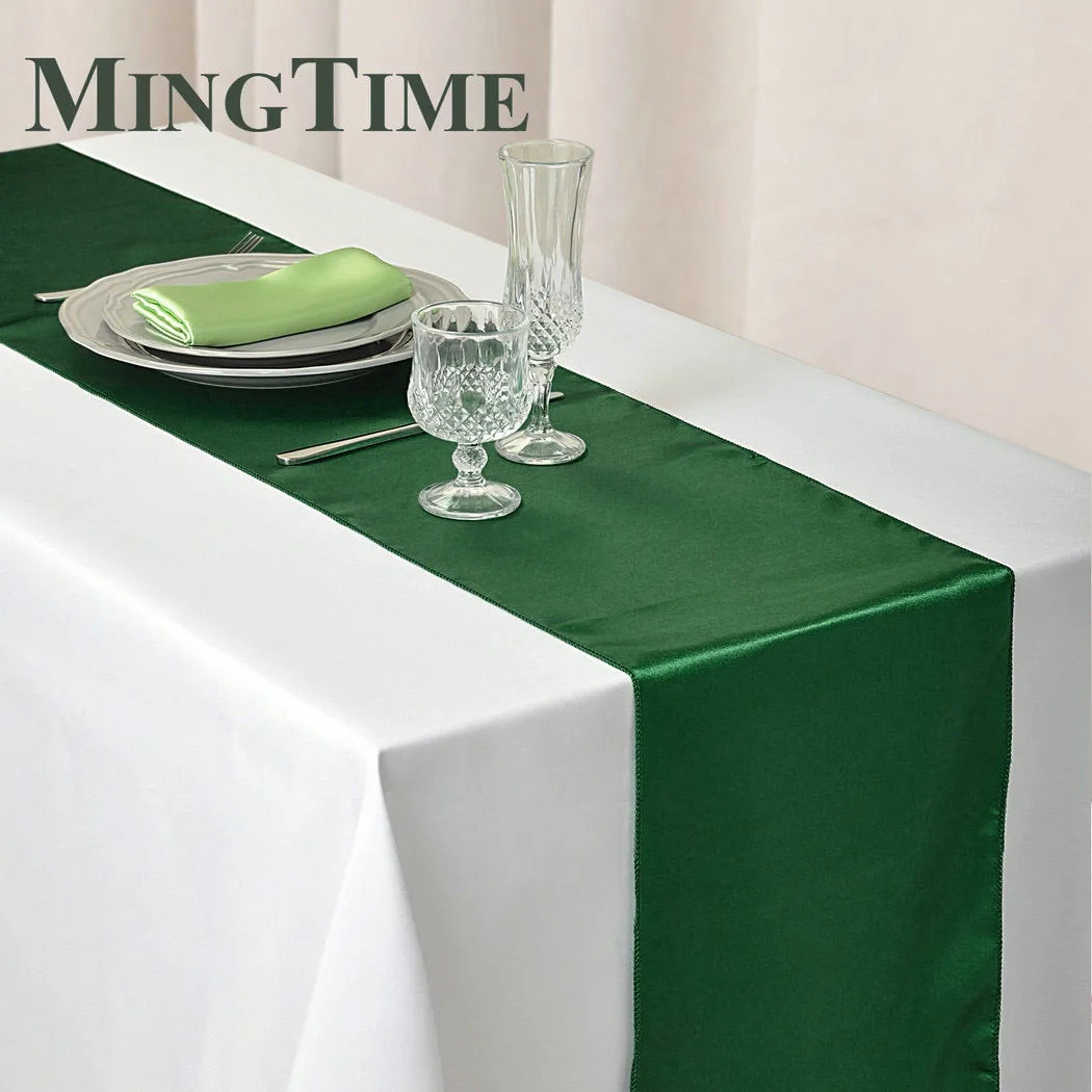 Elegant table setting with a white tablecloth and a green satin runner. It features plates, green napkins, crystal glassware, and cutlery. The background is a light cream color.