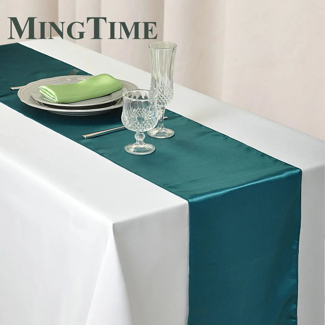 Elegant table setting with teal table runner on a white cloth. Includes crystal glasses, a green napkin on stacked plates, and cutlery. Text "MINGTIME" in the corner.