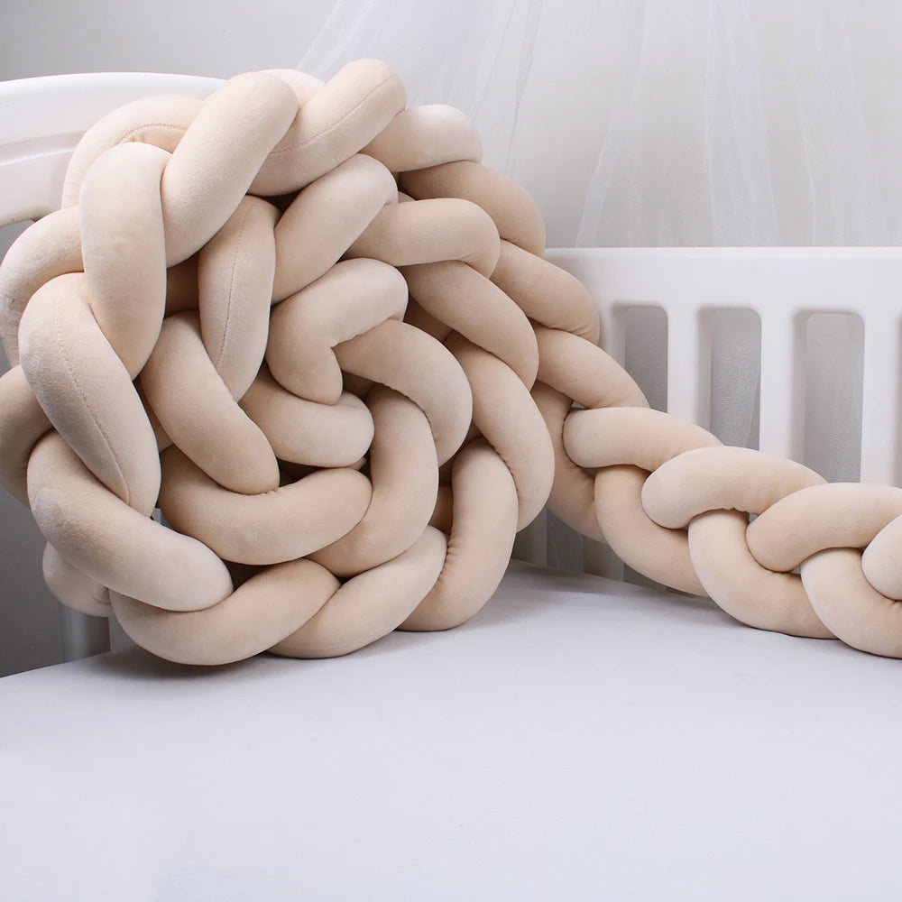 Plush braided cushion in beige draped over the side of a white crib, creating a soft, cozy appearance. The cushion is thick and appears to be made of a velvety fabric.