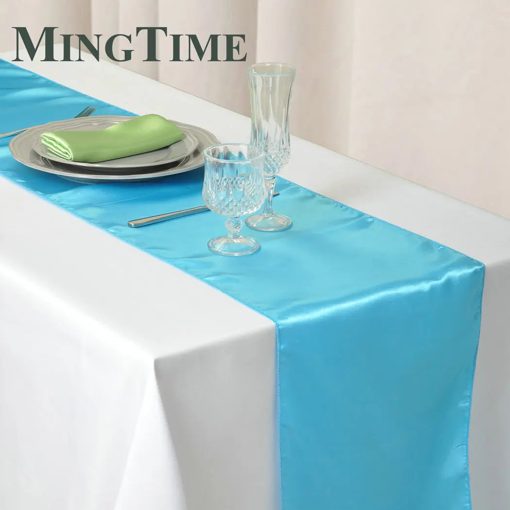 A table set with a bright blue satin runner over a white tablecloth, featuring two elegant glass goblets, stacked gray plates with a green cloth napkin, and cutlery.