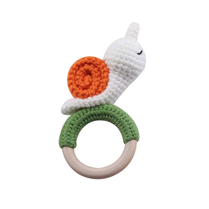 A hand-crocheted snail rattle featuring a white body, orange spiral shell, and a green ring handle with a wooden base, designed for babies.