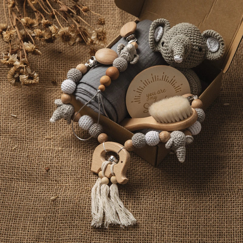 A baby gift set with a crocheted elephant toy, wooden brush, teether, beads, and a soft blanket in a box on burlap. Includes a round card with "you are my sunshine" text.