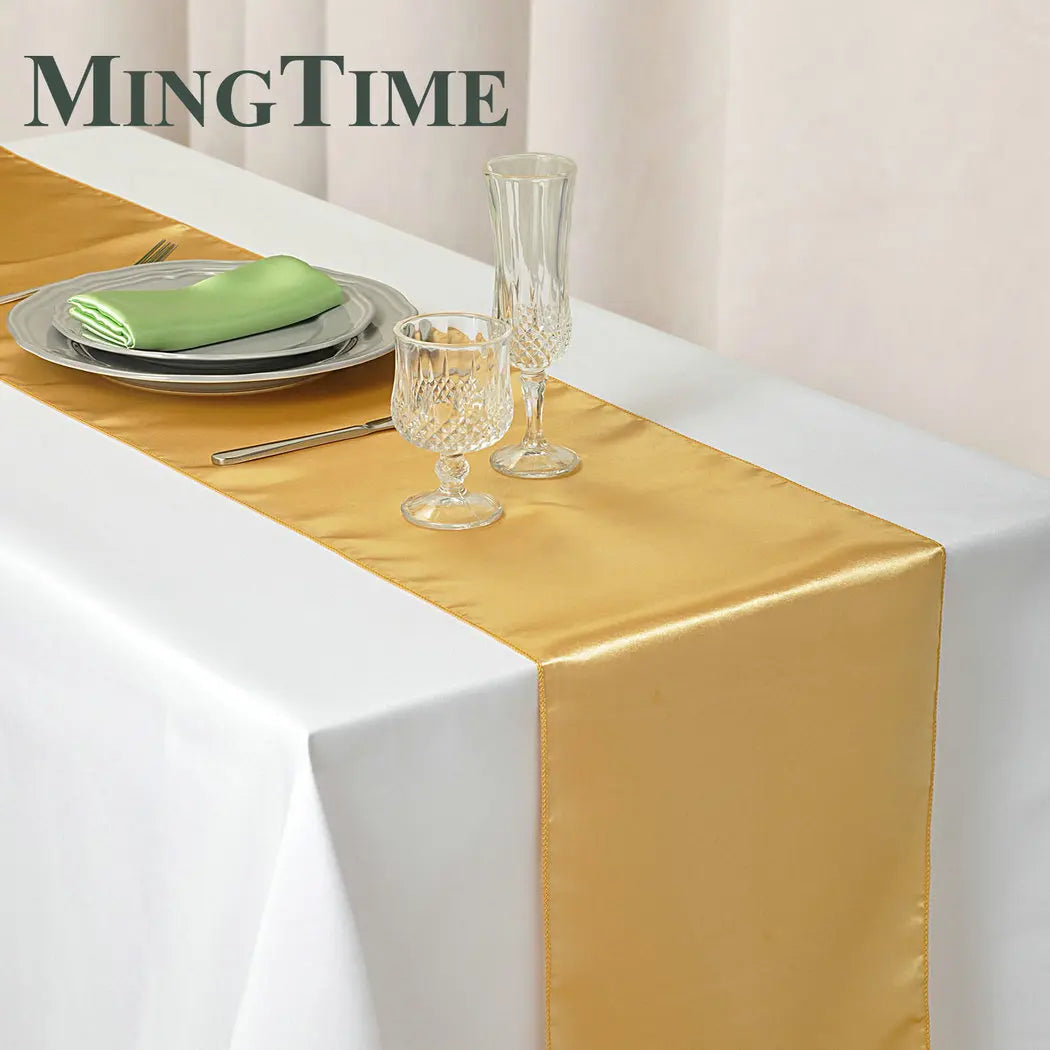 Elegant table setting with a gold satin table runner over a white cloth, featuring a glass goblet and flute, gray plates with a green napkin, and a knife.