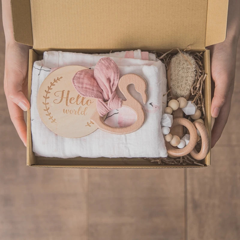 A cardboard box with baby essentials: a wooden "Hello World" sign, pink bow, muslin cloth, natural brush, and wooden teething rings, neatly arranged on a wooden surface.