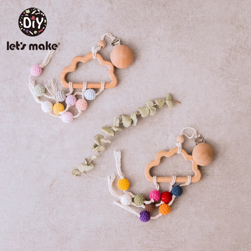 Wooden cloud-shaped baby rattles with multicolored crocheted beads and tassels, set on a light surface with eucalyptus leaves. The DIY "let's make" logo is visible.