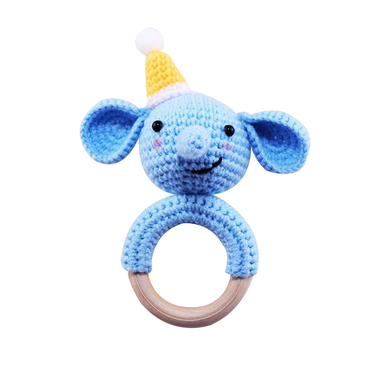 Crocheted blue elephant rattle with a yellow party hat, featuring a wooden ring handle. The elephant has large ears, a smiling face, and pastel pink cheeks.