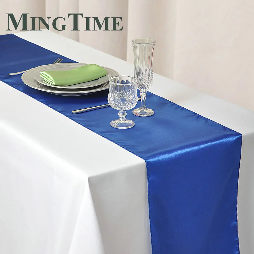 A white tablecloth with a blue satin runner, set with a plate, green napkin, two crystal glasses, and cutlery. The word "MINGTIME" is visible in the corner.