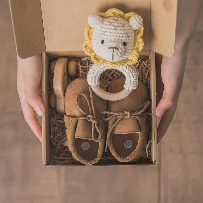 A person holds an open cardboard box containing brown baby shoes with laces and a crocheted lion rattle toy. The shoes rest on straw-like packing material.