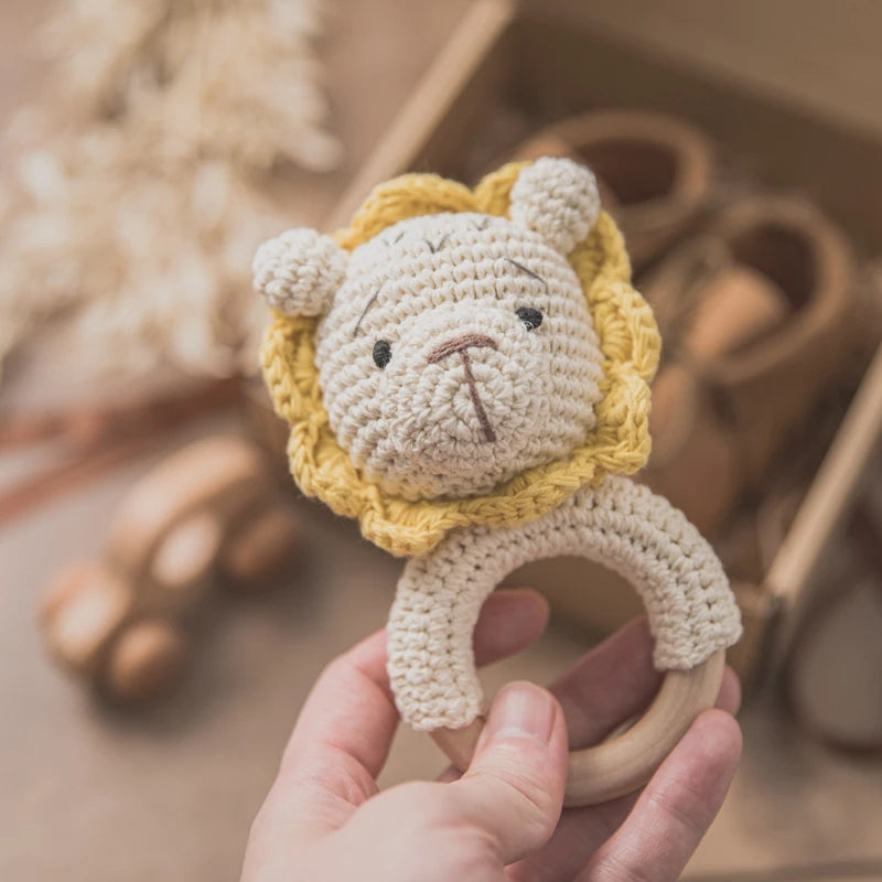 Hand holding a crocheted lion baby rattle with a beige face and yellow mane, featuring a wooden ring handle. Blurred background with wooden toys and shoes in a box.