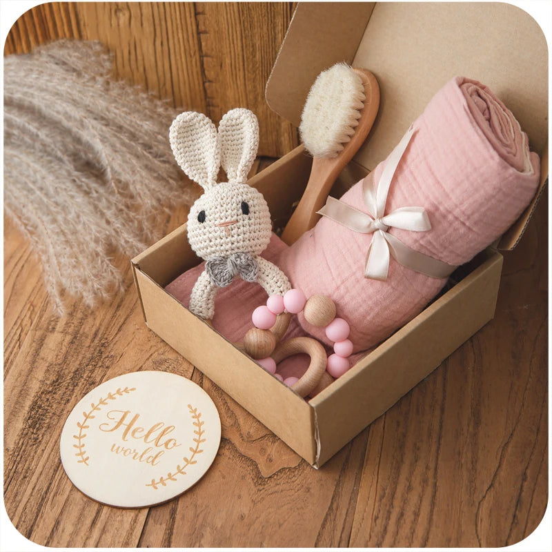 Gift box with a crocheted bunny toy, pink swaddle, wooden brush, and pink beaded teether. A wooden disc with "Hello world" text lies nearby on a wooden surface.