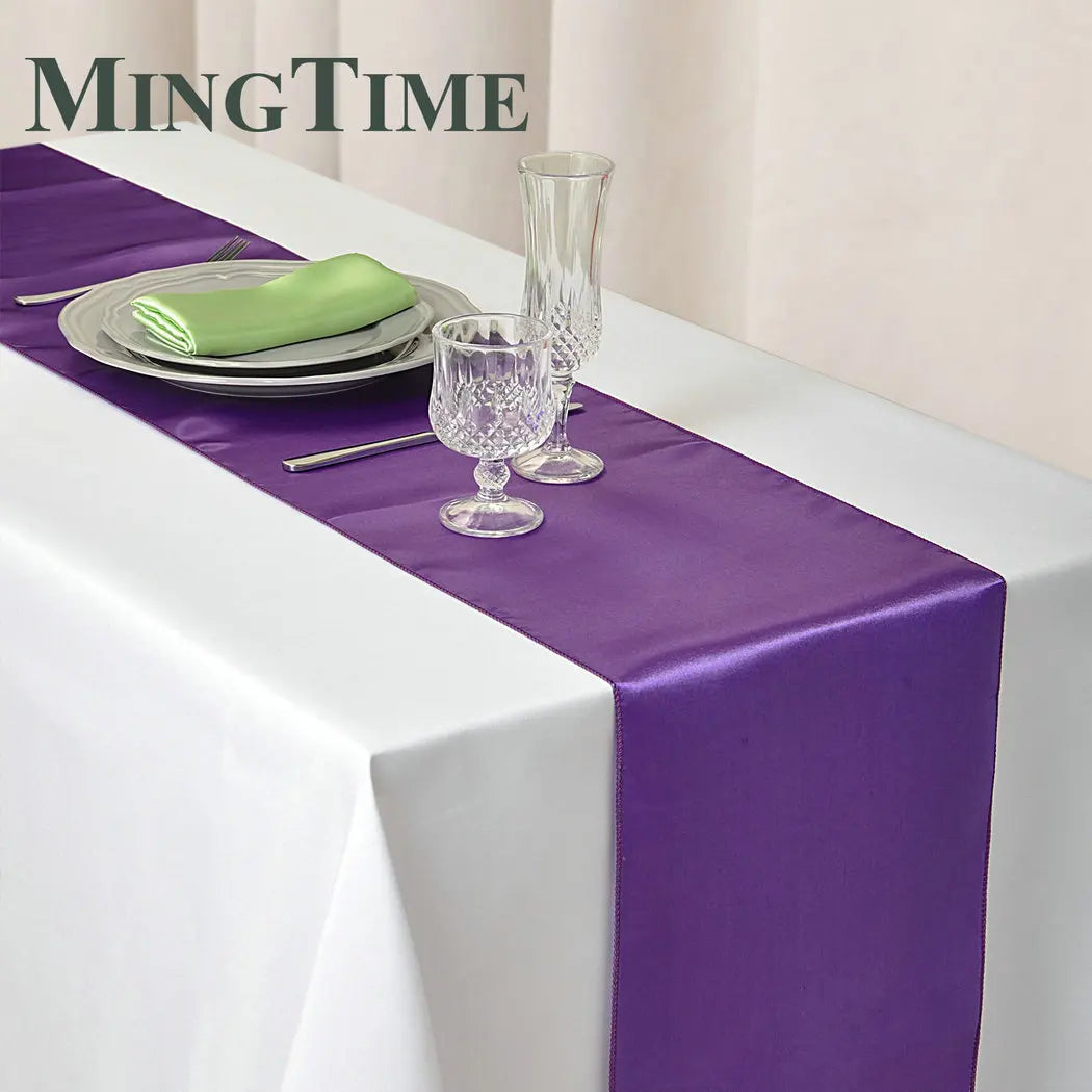 Elegant table setting with a white cloth and purple runner. Crystal glasses, silverware, and a green napkin on a white plate complete the arrangement, branded "MingTime".