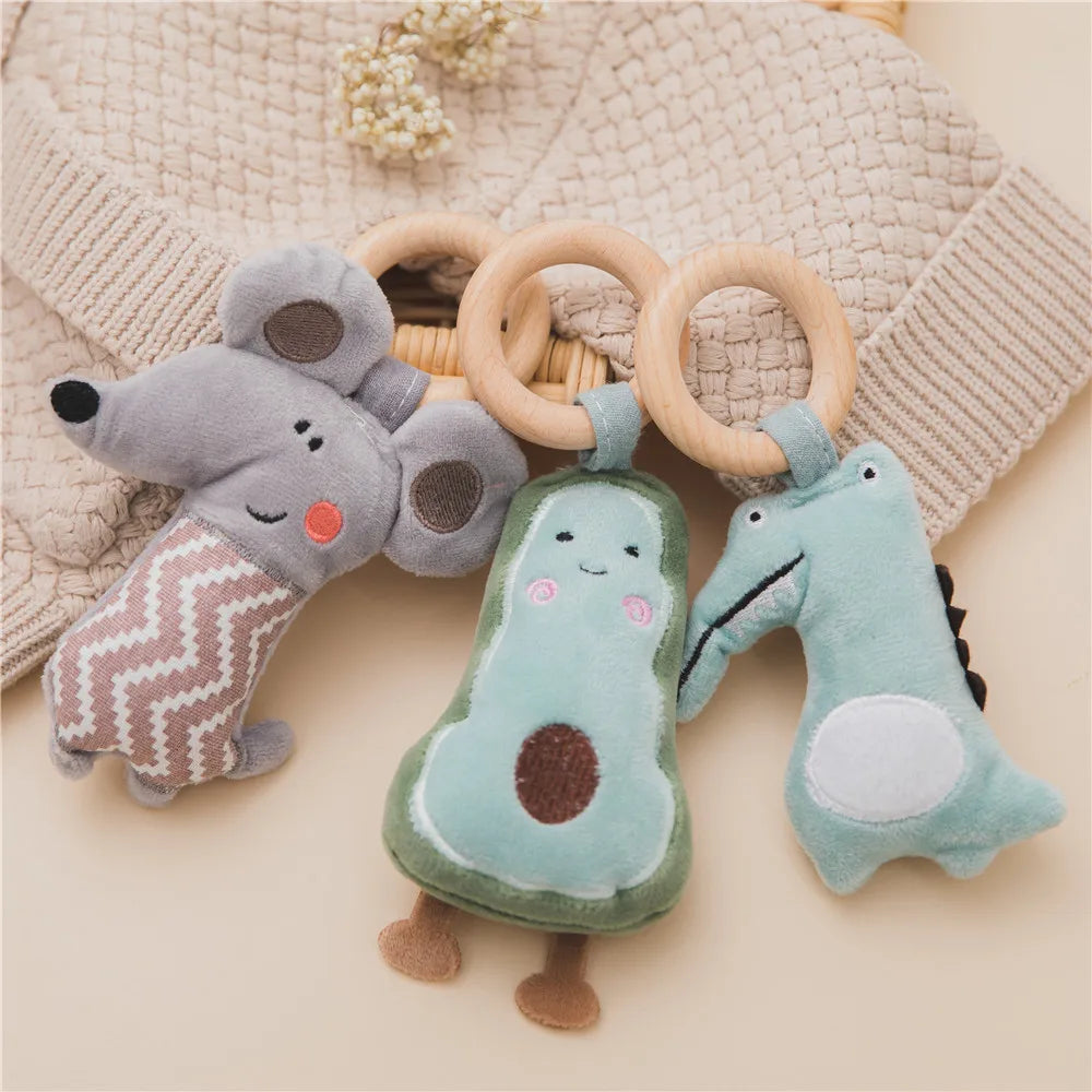 Three plush toys with wooden rings on a textured beige sweater: a gray mouse, a smiling avocado, and a teal crocodile, arranged under small dried flowers.