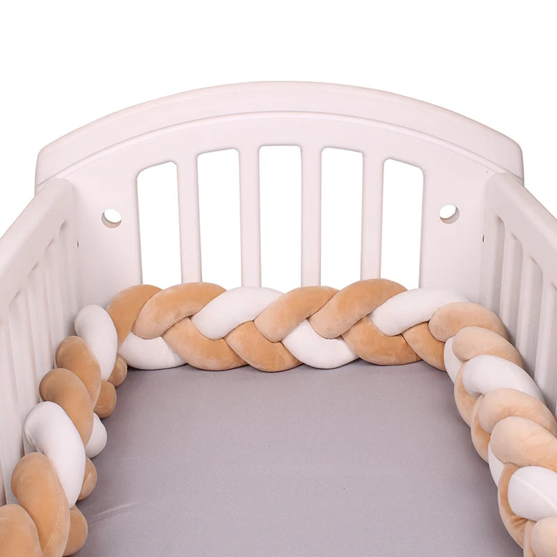 White baby crib with a soft braided bumper in beige and white placed around the inside edges. The mattress is covered with a light gray sheet.
