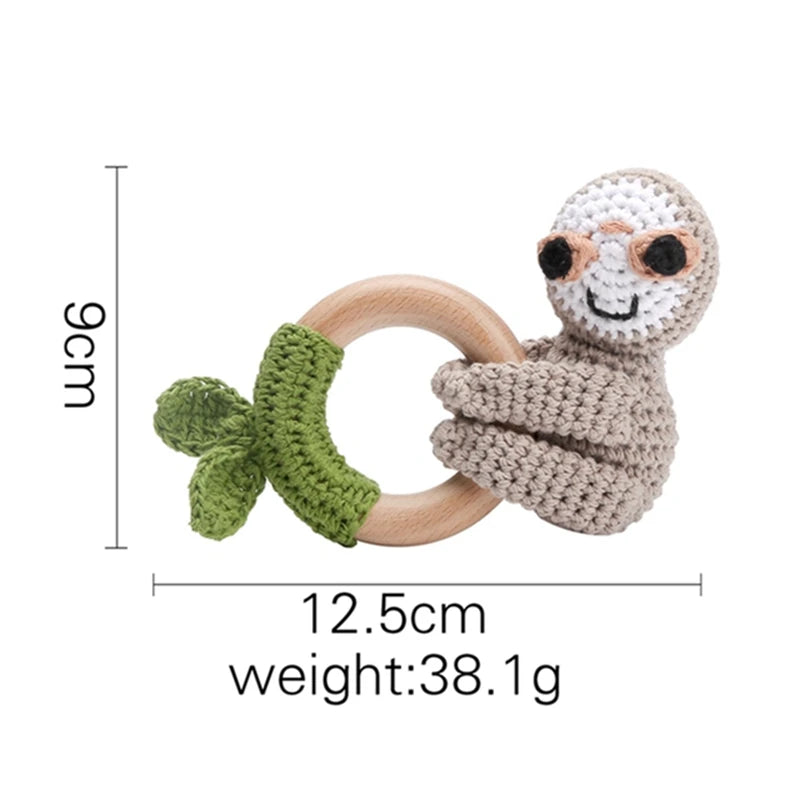 Crocheted sloth baby rattle with a wooden ring, green leaf detail, and smiling face. Measures 9cm x 12.5cm, weighing 38.1g. Perfect for sensory play.