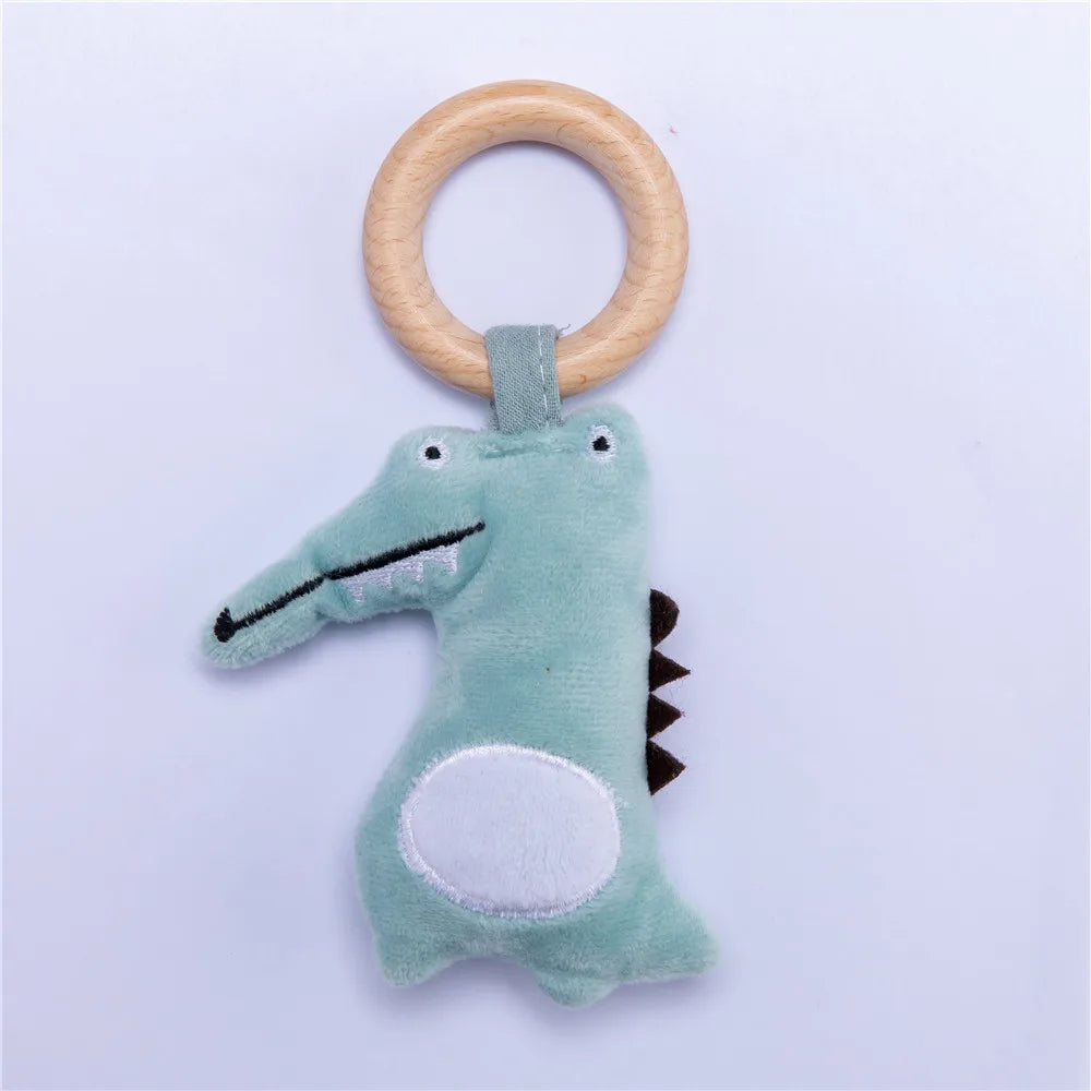 Soft plush crocodile toy with a wooden ring handle, featuring a light green body, white belly, embroidered smile, and small black spikes on its back, set against a plain background.