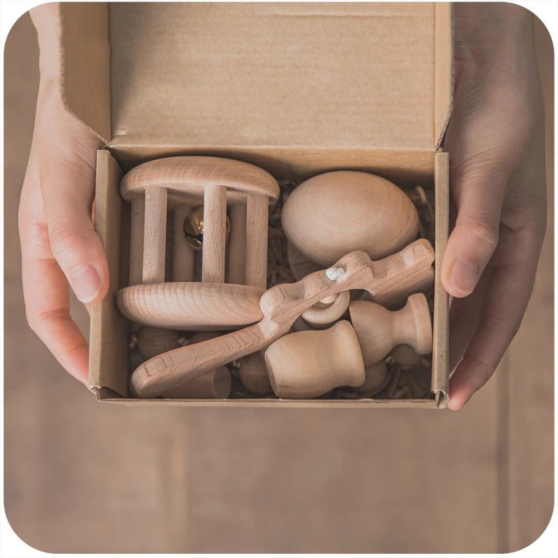Hands hold a cardboard box containing wooden toys including a rattle, egg, and stacking figures on a neutral background.