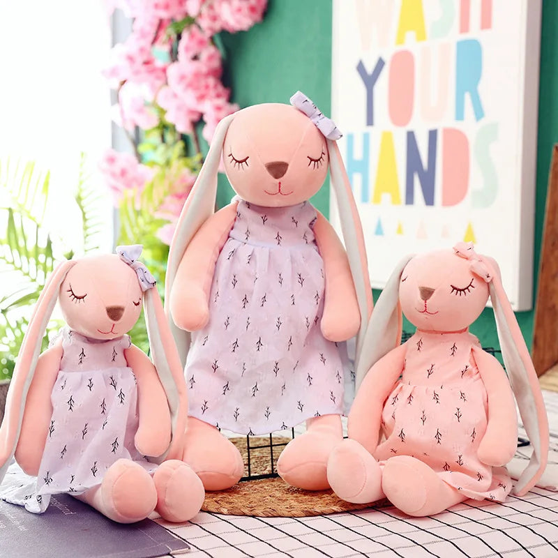 Three pink bunny plushies with long ears and floral dresses sit on a table. A colorful "Wash Your Hands" sign and pink flowers are in the background.