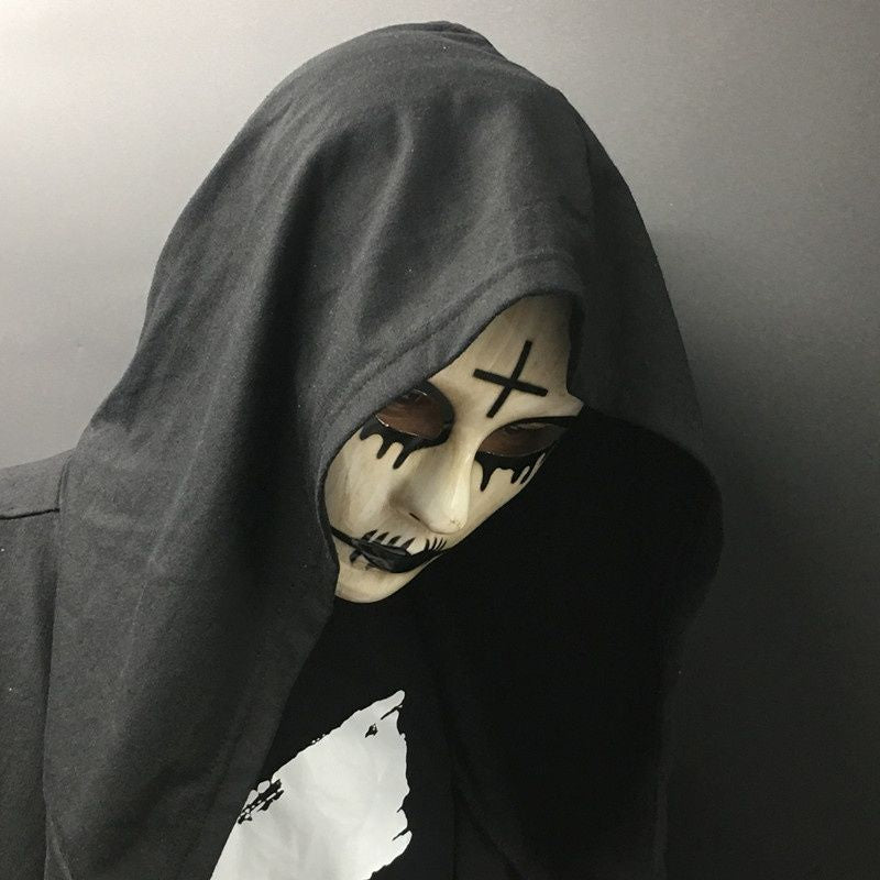 A person wearing a black hooded cloak and eerie white face paint with black accents, including an X on the forehead, set against a plain dark background.