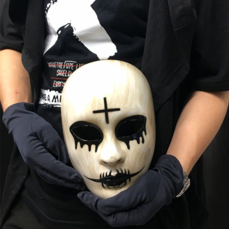 Person wearing black gloves holds a white mask with black cross on forehead, dripping eye designs, and stitched mouth pattern, against a backdrop of a black shirt with white graphics.