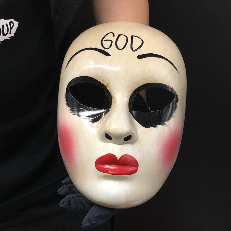 A person holds a white mask with exaggerated makeup, red lips, and the word "GOD" written on the forehead. The mask features black paint around the eyes and rosy cheeks.