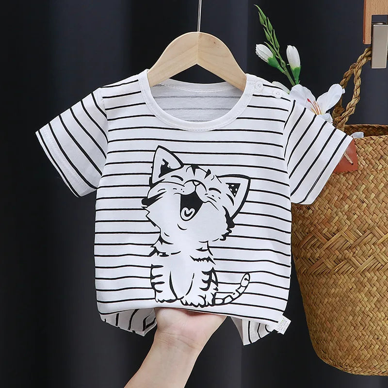 A white baby t-shirt with black horizontal stripes and a cute, smiling cartoon cat design, held on a hanger. A woven basket and white flowers are visible in the background.