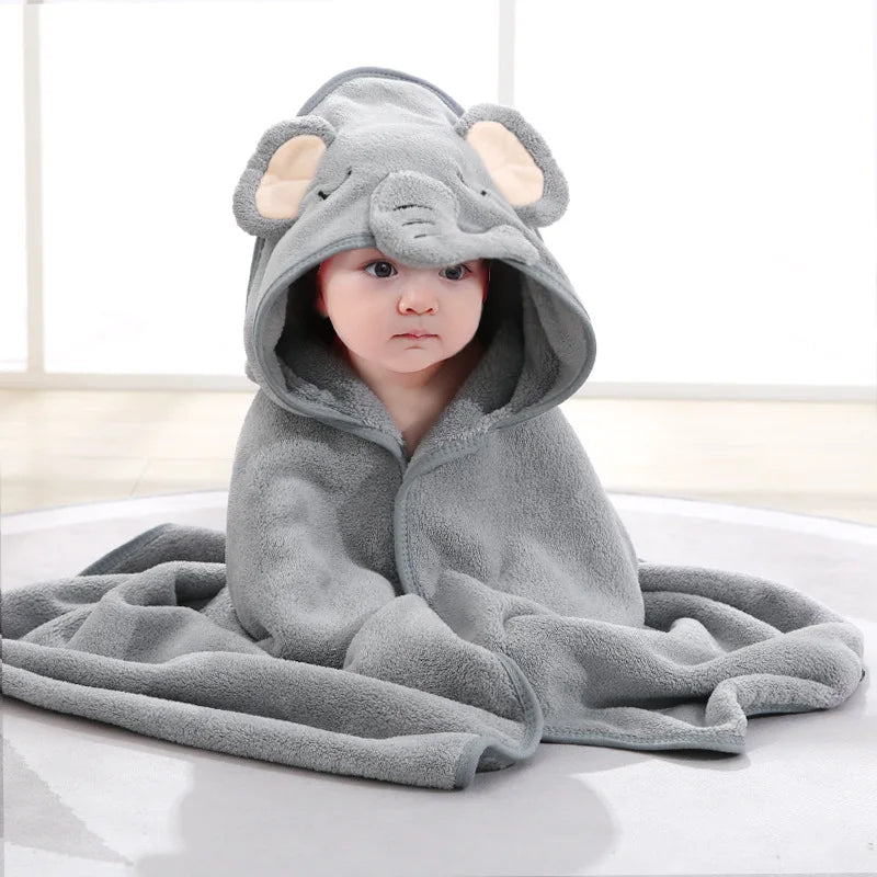 A baby sits on a light gray rug wearing a soft, gray elephant-themed hooded blanket with floppy ears. The room is bright with a pale floor and a large window.