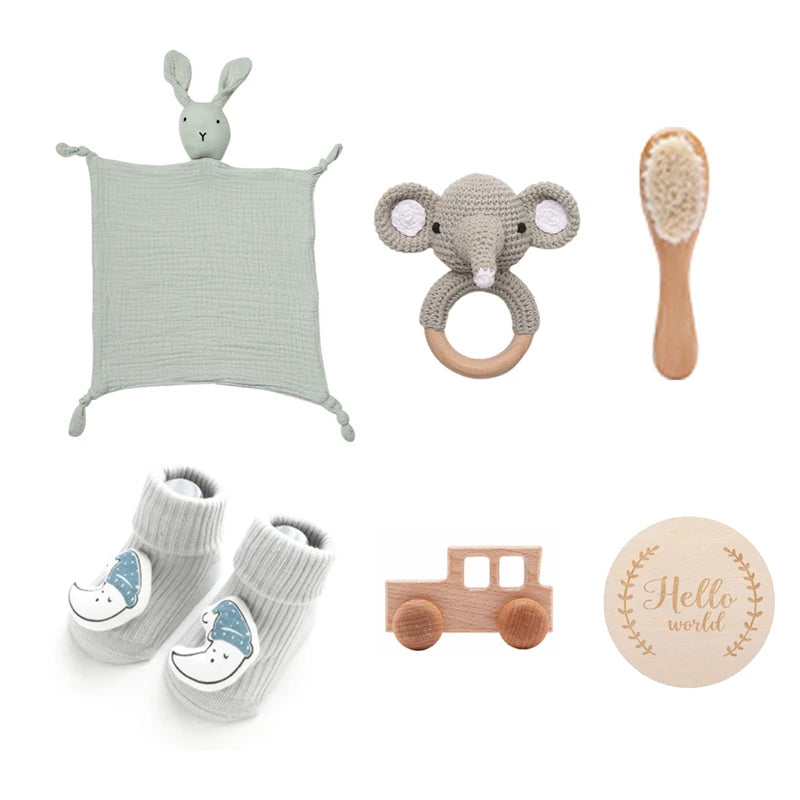 A collection of baby items: a soft green rabbit blankie, a gray elephant rattle, a wooden brush, gray socks with moon designs, a wooden train, and a “Hello world” sign.