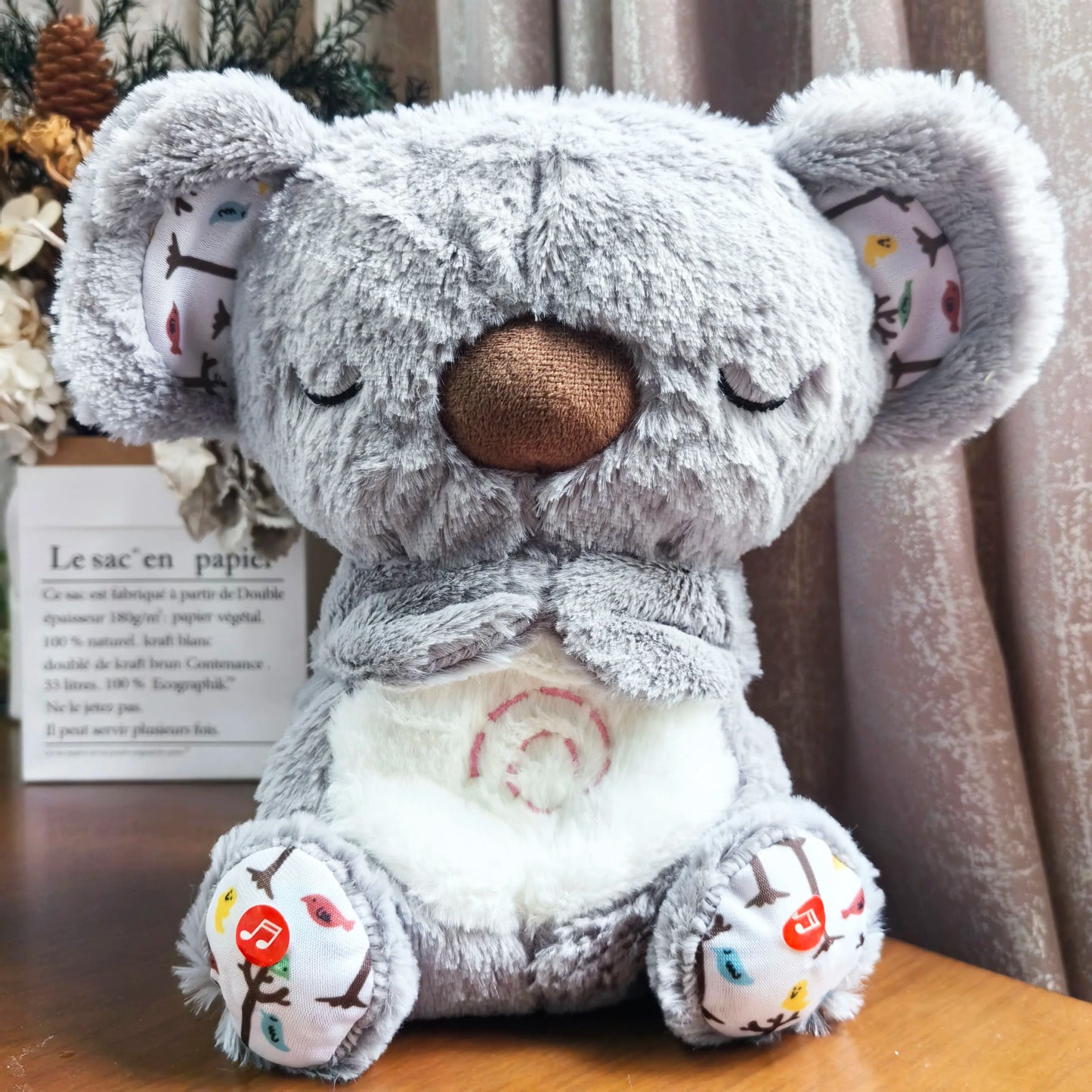 A plush gray koala toy with closed eyes and a brown nose sits on a wooden surface. Its ears and feet have colorful patterns, and a music icon is on one foot. Background includes dried flowers.