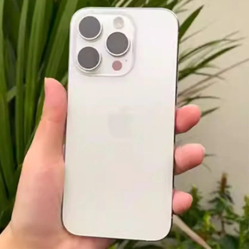 A person holding a white smartphone with three large camera lenses on the back, set against a background of green plants and beige wall.