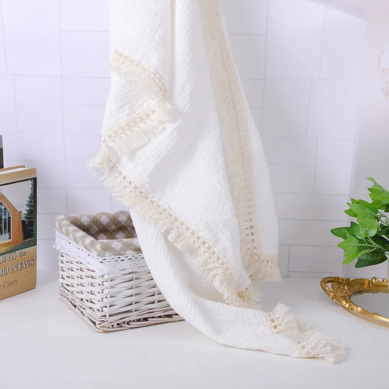 A white textured towel with lace edging is draped over a wicker basket. A book and a small green plant are nearby on a white surface against a tiled wall.