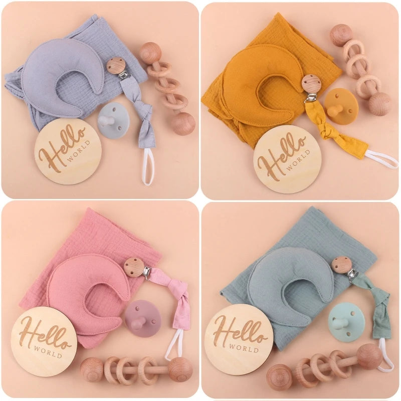 Four baby gift sets, each with a cloth, wooden teether, pacifier, and "Hello World" plaque. Colors: blue, mustard, pink, and mint green on a beige background.
