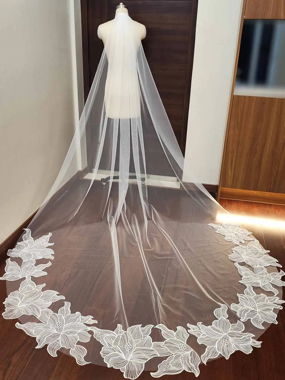 A mannequin displays a long, sheer wedding veil with intricate white floral embroidery along the edge, draped elegantly in a room with wooden flooring and furniture.