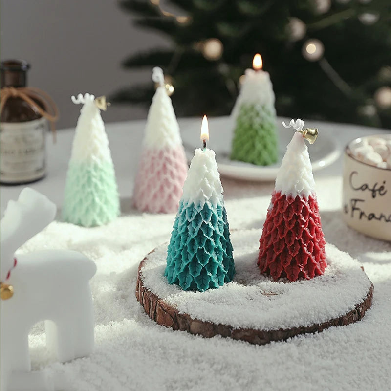 Festive tree-shaped candles in red, green, blue, and pink, placed on snow-like powder, with blurred background of a Christmas tree and holiday decor. Two candles are lit.