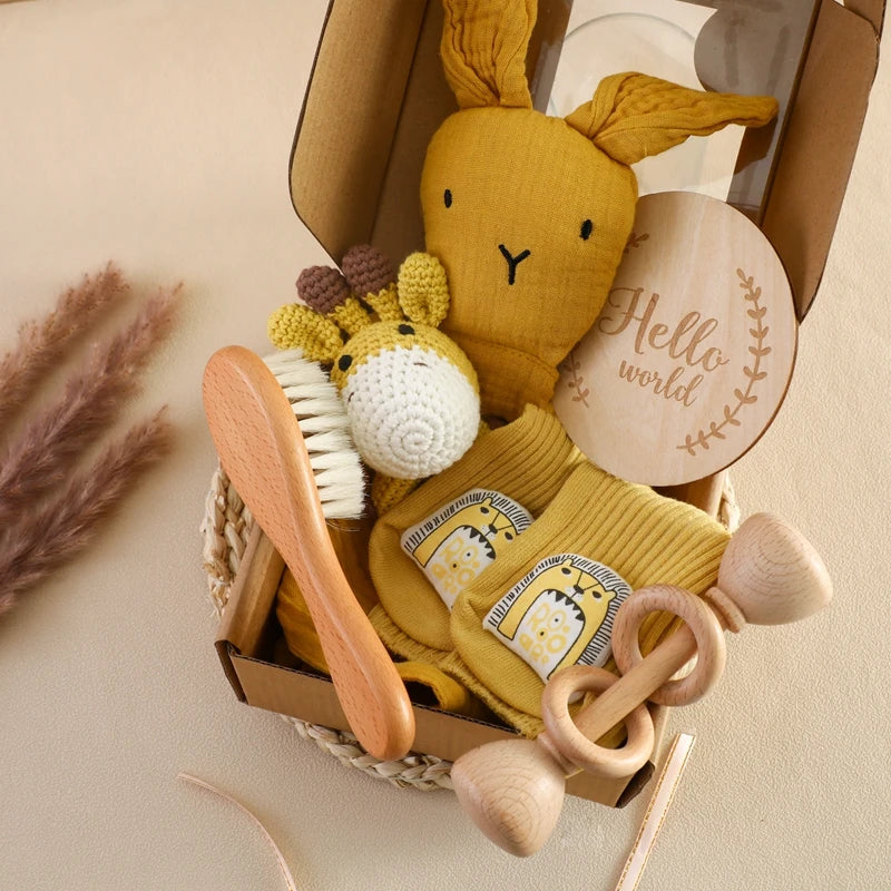 A cozy baby gift box with a mustard yellow stuffed rabbit, a crocheted giraffe, wooden brush and rattle, yellow socks with giraffe print, and a "Hello world" plaque.