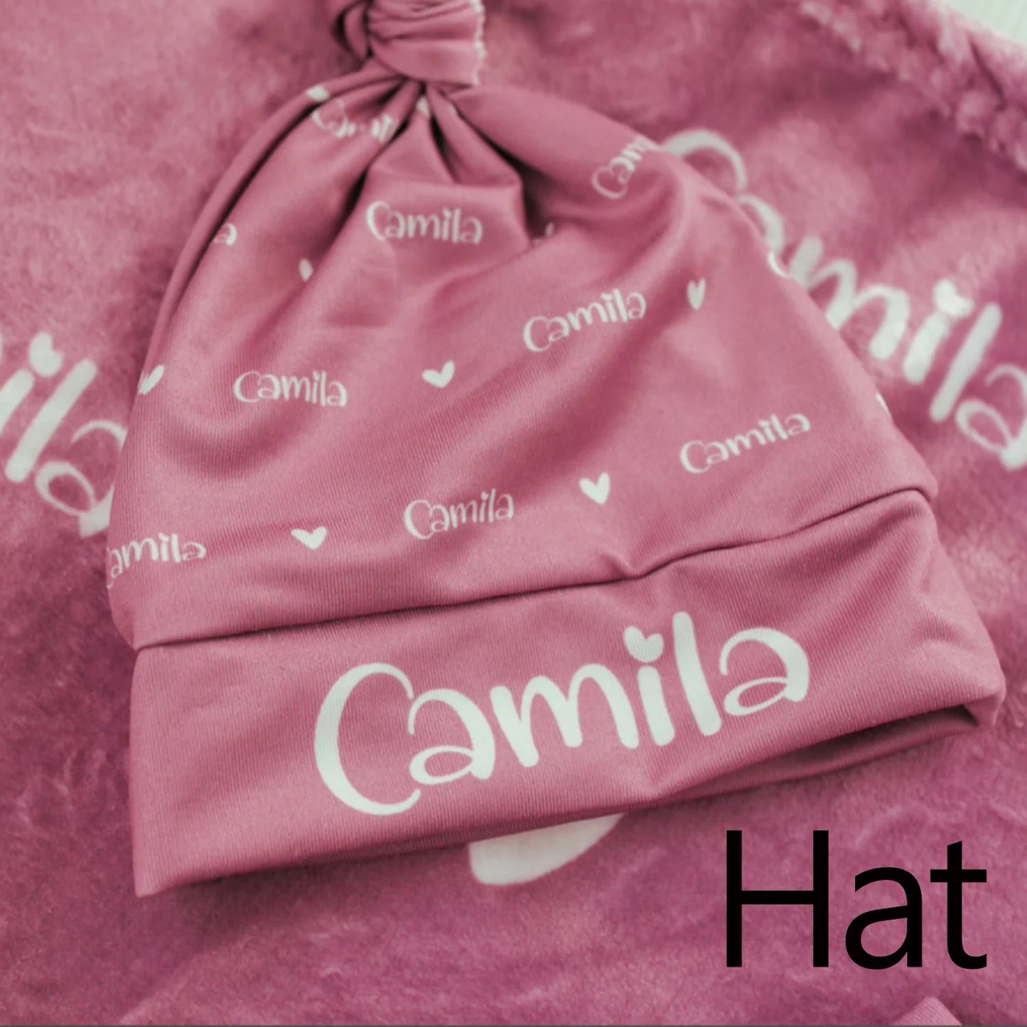 Pink baby hat with "Camila" and small hearts printed on it, laid on a matching pink fabric, with "Hat" text overlay in the bottom right corner.