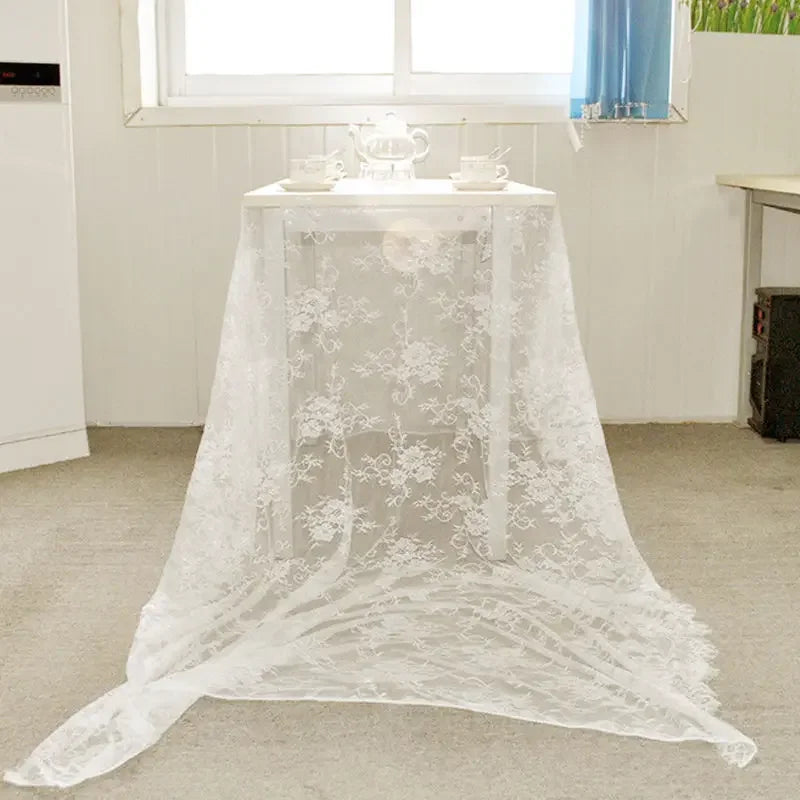 A small table with a delicate lace tablecloth, set for tea with a teapot and cups, near a window with blue curtains, in a softly lit room.