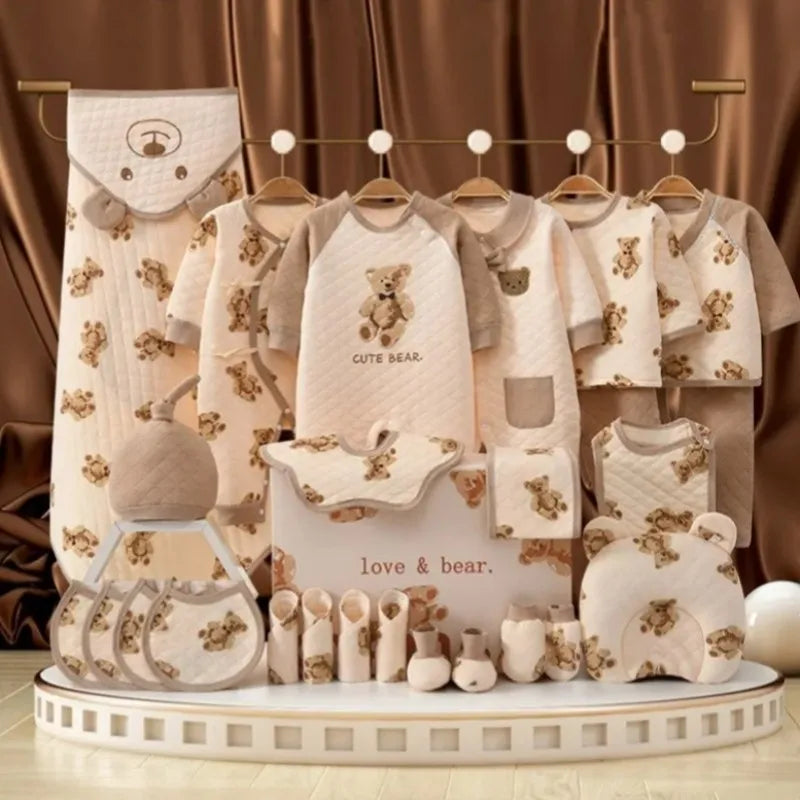 A display of beige and brown baby clothing and accessories, all featuring teddy bear patterns. Items include onesies, bibs, hats, mittens, and a pillow, set against a brown curtain.