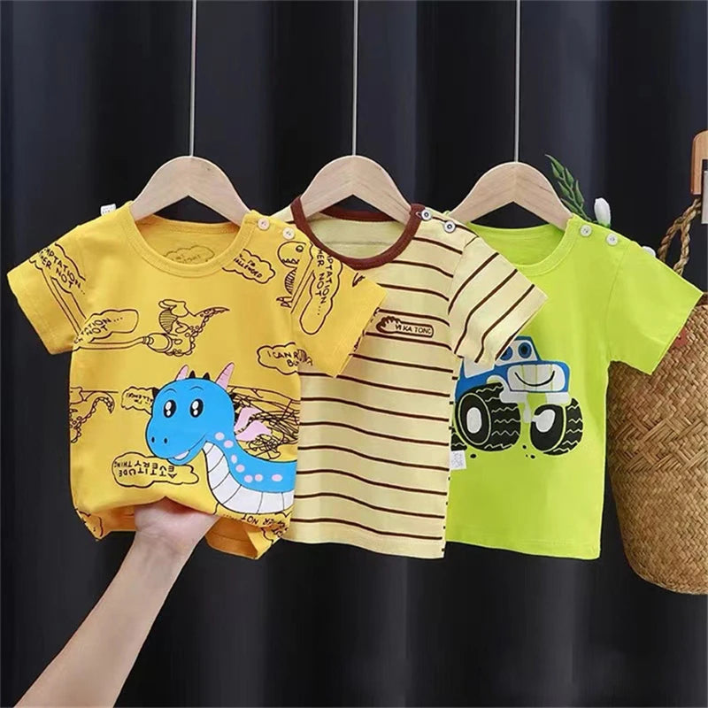 Three colorful baby shirts on hangers: a yellow one with a blue dinosaur, a yellow striped one, and a bright green one with a truck design, set against a dark backdrop.