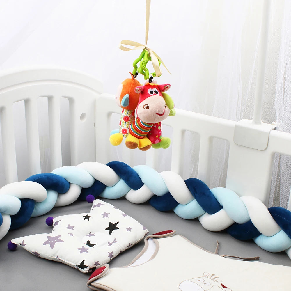 Colorful plush toys hang above a white crib, which is lined with a braided blue and white bumper. A star-patterned pillow and soft garment are placed inside.