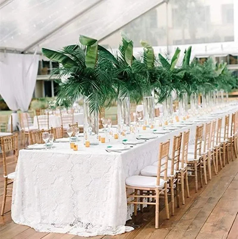 Elegant outdoor dining setup under a tent, featuring a long table with white lace cloth, rows of wooden chairs, and tall glass vases with lush green palm leaves as centerpieces.