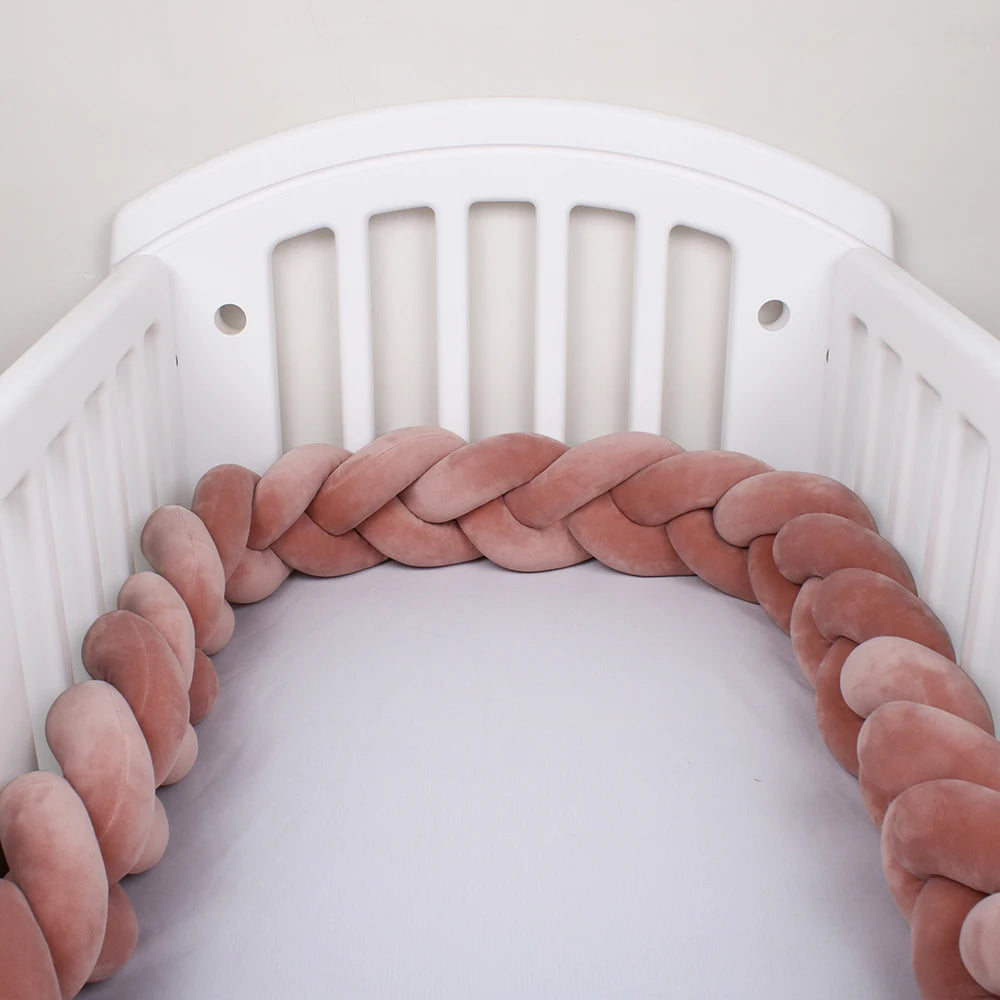 A white crib with a soft, pink braided cushion lining the inside edges, creating a cozy and protective barrier on the mattress.