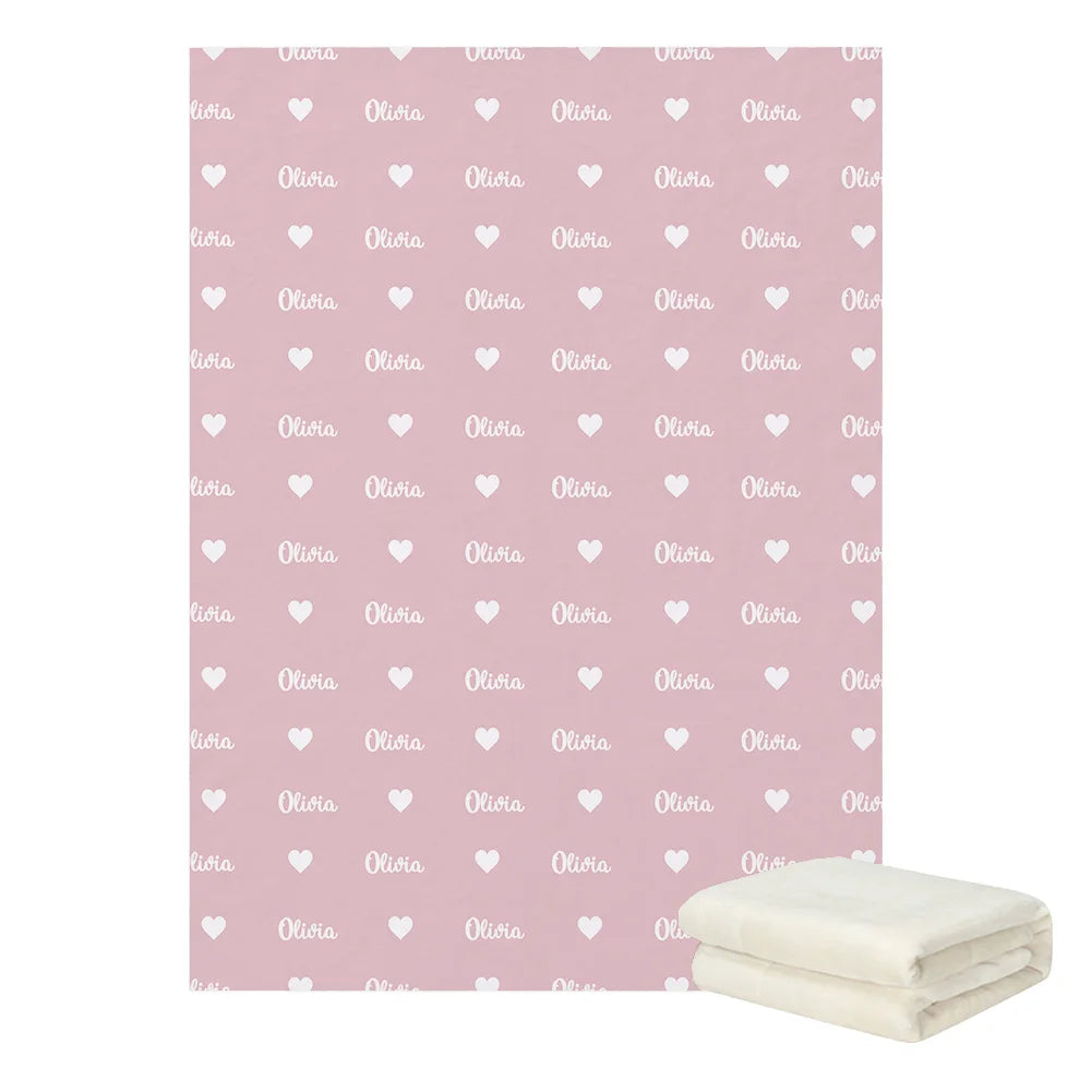 Pink blanket with repeated white heart and "Olivia" patterns. A folded cream-colored blanket is placed beside it, indicating a matching set or additional piece.