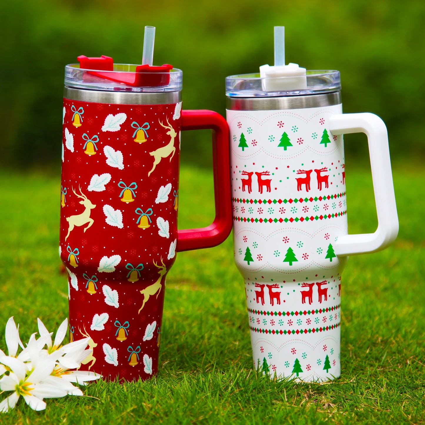 Two festive travel mugs on grass; one red with reindeer, bells, leaves, and snowflakes, and another white with red reindeer, green trees, and geometric patterns, both with lids and straws.