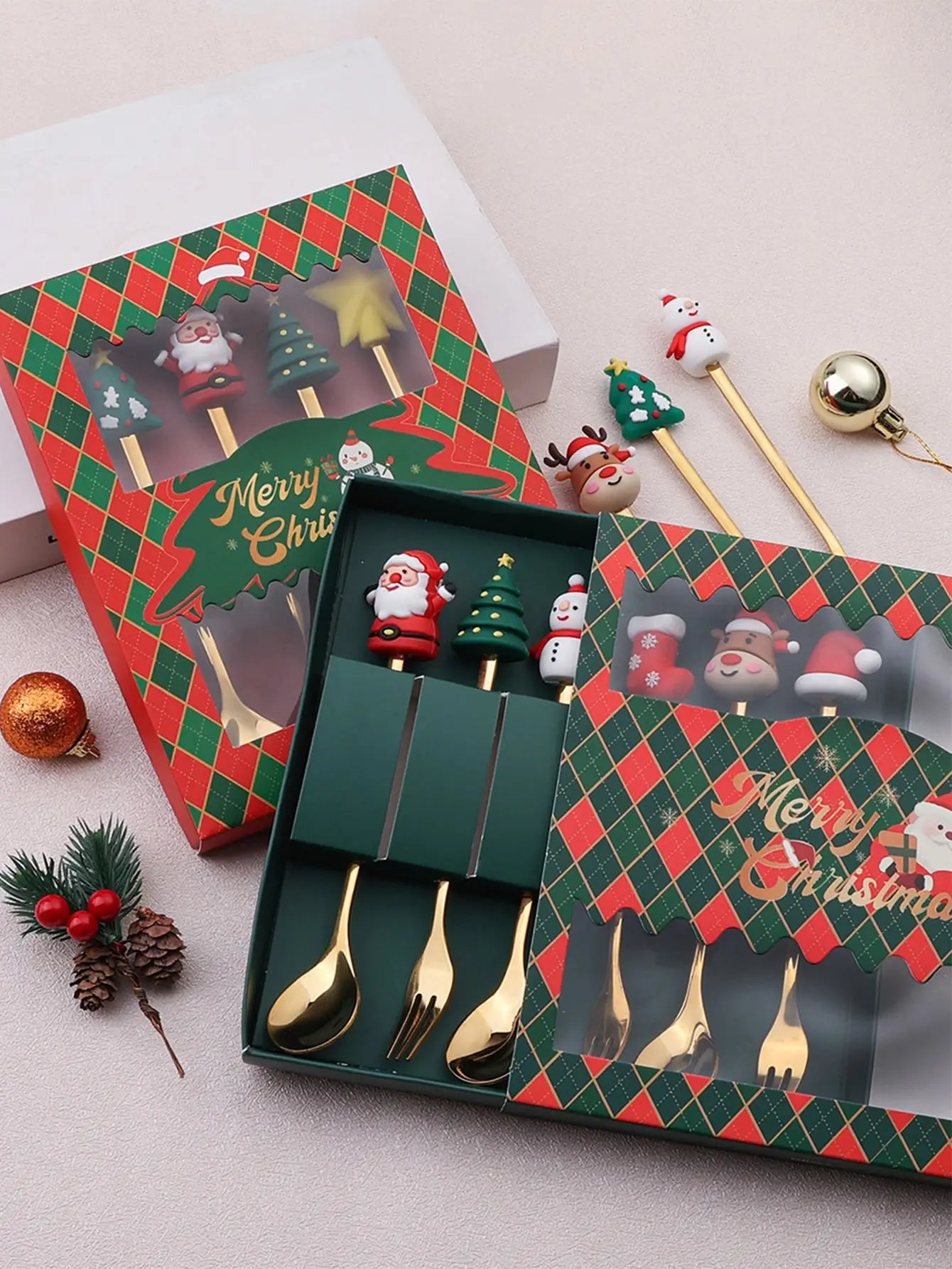 Festive cutlery set in a Christmas-themed box with golden spoon and fork. Includes decorative sticks with Santa, snowman, and tree designs. Accompanied by ornaments and pine cones.