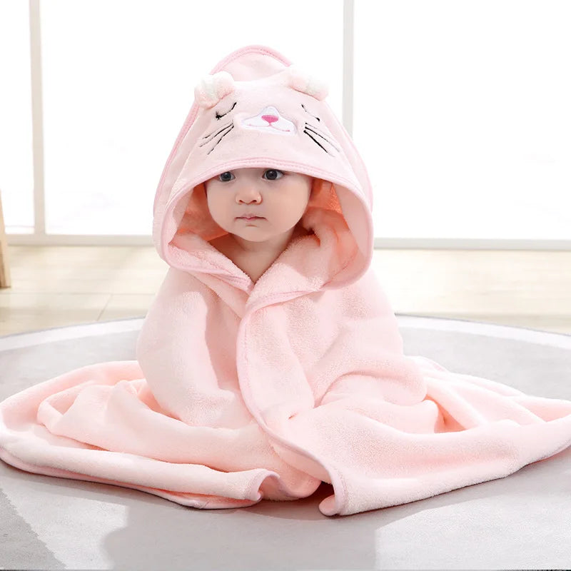 Baby wrapped in a soft pink blanket with a hood featuring a cute animal face sits on a light gray rug. The room is softly lit and has a bright background.